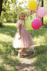 a small beautiful blonde girl in a pink dress with balloons in her hands is walking through the park