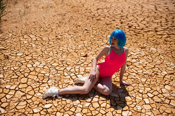 Beautiful young model girl with short blue hair and pink bathing suit  posing in the desert. Cracks in the sand 