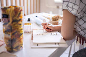 Smiling young beautiful business female taking notes in paper diary at modern Nordic style kitchen