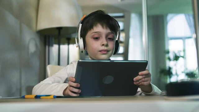 Curious Little Schoolboy Sitting At The Table Listening To Music And Playing Games On The Tablet Watching Video Internet Education Concept