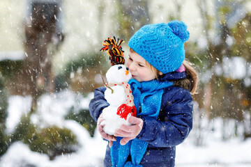 Cute little toddler girl making mini snowman and eating carrot nose. Adorable healthy happy child playing and having fun with snow, outdoors on cold day. Active leisure with children in winter