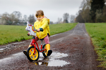 Little toddler girl wearing yellow rain gum boots, running with balance bike during sleet. Happy...