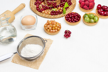 Flour in sieve, egg and rolling pin, bottle of water. Tartlets with berries. Red currants on wicker plate.