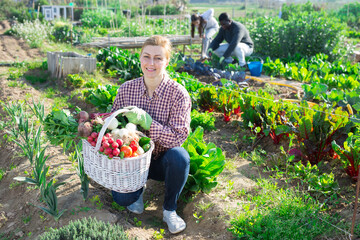 Positive woman farmer holding wicker basket full of freshly harvested vegetables on a farm field on a warm spring day