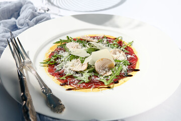 Beef carpaccio with mushrooms and parmesan cheese. Fancy dinning with beef carpaccio on white table with simple contemporary decor. Sunlight and harsh shadow still life. Beef carpaccio appetizer dish.