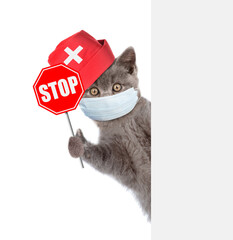 Kitten wearing doctors cap and medical protective mask shows stop sign and look from behinde empty banner. Isolated on white background