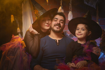 Mom, dad and daughter in costumes for holiday of halloween in dark decoration and with nice yellow light. Funny nice family during carnival in room. Mother, father and girl during funny photo shoot