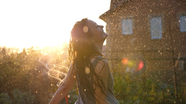 Joyful brunette girl enjoys her evening in the countryside by dancing in the rain. Stunning golden sun rays shine on playful young woman spinning and enjoying a spring shower. CLOSE UP, LENS FLARE