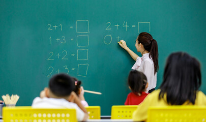 Portrait shot of Asian beautiful female mathematic tutor standing smiling holding chalk writing elementary school math equation questions on chalkboard while group of boy and girls students studying
