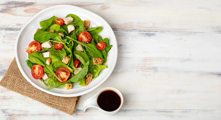 Fresh spinach salad with tomato, small pieces of herb roasted chicken in white ceramic dish on brown sackcloth, together with balsamic vinegar on white wooded table. Concept for healthy