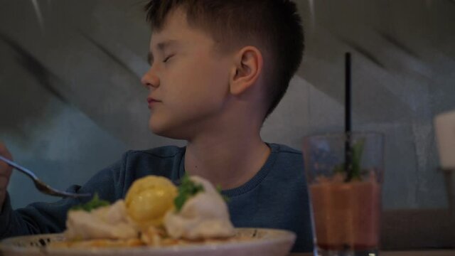 a boy eats dessert in a cafe. waffles with cream and smoothies in a glass. a smile on his face. he winces at the cold