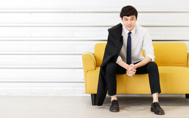 Fototapeta premium Horizontal portrait shot of young adult Asian handsome man in a white shirt with a suit on the shoulder sitting on the sofa and looking at the camera with hand to chin gesture posing in the studio