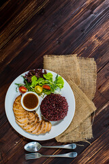 Top view flat lay shot of a sliced oven-roasted black pepper chicken with riceberry rice, sesame sauce, and salad side dish on the wooden table with spoon and fork. Concept of clean food, healthy