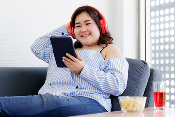 Asian young happy pretty friendly fat plus size woman sit smiling relaxing on gray fabric couch listening to music from big red headphones use playlist from black tablet on hand in living room