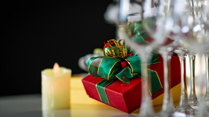 Close up shot of green and gold ribbon bow tied red luxury precious present gift box placed on desk behind empty tall wine glasses and candles in blurred foreground celebrate anniversary dinner night