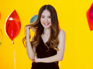 Asian young sexy beautiful long hair girl in dress standing smiling posing happily in front of colorful star and round shape helium air balloons on yellow background in surprise party