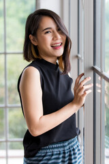 Horizontal portrait shot of a cute smiling Asian woman with a stylish straight hairstyle in black clothes standing, touching the windows, and looking at camera. Concept of happiness at home