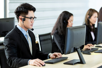 Cheerful Asian man in headset smile with cheerful and happy while working on computer at desk with female coworkers in help desk office. Sevice mind and ready  for marketing support concept