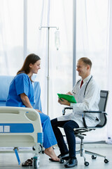 Obraz premium Woman caucasian patient sitting on bed and caucasian doctor sitting on chair are talking, both are smiling look happy. Doctor man carrying stethoscope on neck and hold writing on note in office room