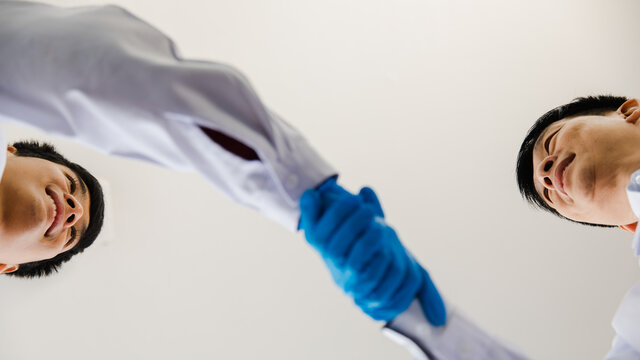 From bottom view close-up focus to 2 hands are handshakes by 2 adults man who are scientist wearing white uniform and blue glove