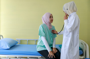 Young female Muslim doctor checking Muslim patient's heart by stethoscope at hospital while sitting...