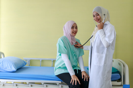 Young female Muslim doctor checking Muslim patient's heart by stethoscope at hospital while sitting on hospital bed