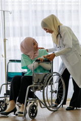 Fototapeta premium Young female muslim doctor talking and cheering up young sick muslim pateint sitting on wheelchair in hospital