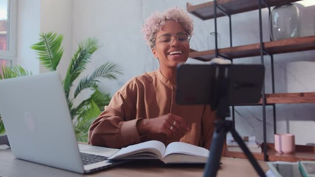 Happy african american woman turns on cell phone camera to record video. Beautiful young woman sitting with book and laptop and going to record useful video for her blog using phone on tripod.