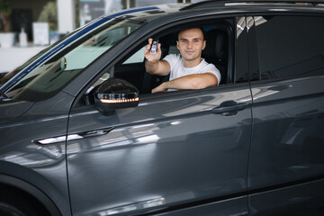 Fototapeta na wymiar Man sitting in his new car in car showroom. Portrait of handsome man in car hold keys in hands