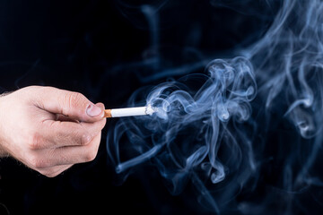 Black background. the hand of a man with roughened skin holds a cigarette. blue smoke is coming...