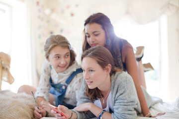 Three teenage girls using smart phone together while lying on bed in bedroom