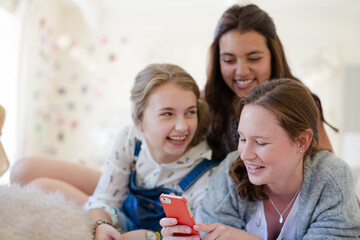 Three teenage girls using smart phone together while lying on bed in bedroom