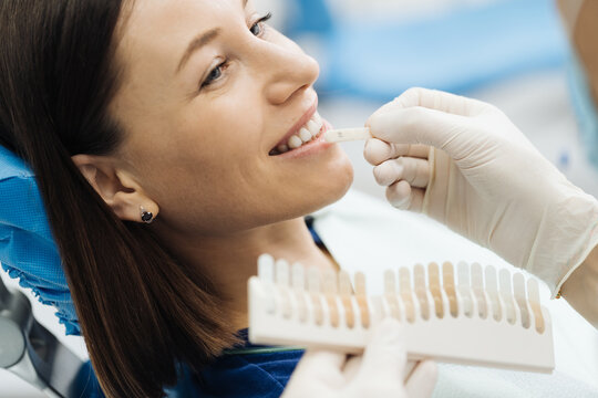 Close up portrait of beautiful young lady sitting in dental chair while stomatologist hands in sterile gloves holding tooth samples. She is smiling