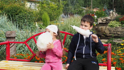 Happy cute boy and girl eating cotton candy floss in the park near the fountain sitting on a bench....