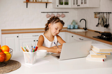 Obraz na płótnie Canvas Little pretty girl with two ponytails sitting at kitchen at table with exercise book and laptop doing her school lessons because of lockdown quarantine. Home family education concept.