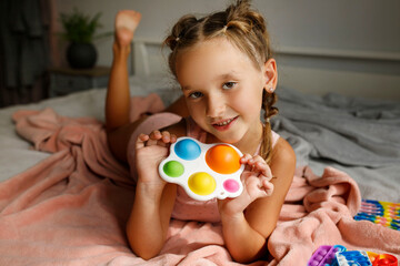 Pretty little girl in pink pajamas laying in her bad at daytime and playing antistress multicolored...