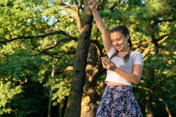 Happy girl mobile. Euphoric screaming young girl holding smartphone showing winner gesture at sunny day in park. Modern technologies and communication.