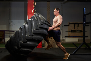 a man training in a gym with a naked inflated torso lifts a huge car tire with great physical effort and holds it on his knee. strength exercises in the gym