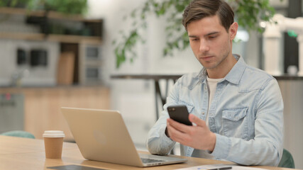Young Man with Laptop using Smartphone at Work