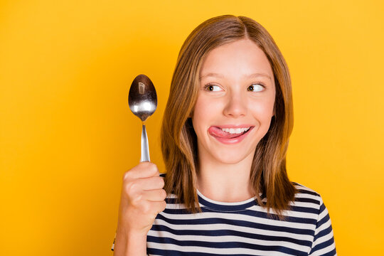 Portrait of positive pupil look hand hold spoon tongue lick teeth have good mood isolated on yellow color background