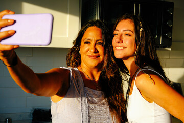 Mature mother taking selfie with smiling daughter in kitchen at home