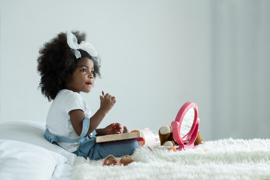 Little African girl playing makeup with mother's cosmetics. Adorable kid painting her lips with red lipstick and looking in the mirror with joy in bedroom at home