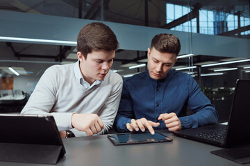 Young businessmen sitting in office and checking tablet with company report, laptops standing beside. Managers discussing progress, Agile board arranged on glass wall behind them. Concept of planning