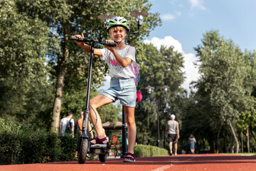 Profile view portrait of cute blond little caucasian school girl wear helmet enjoy having fun riding electric scooter city street park outdoors on sunny day. Healthy sport children activities outside