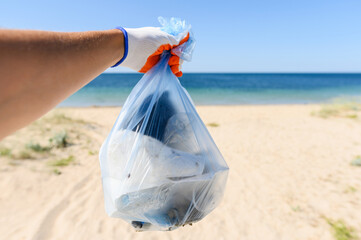 A full bag of trash in a man's hand against the backdrop of a clean beach and sea. Garbage collection concept. Selective focusing.