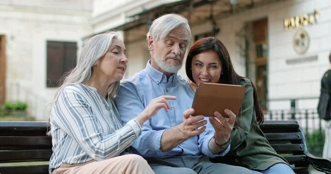 My modern parents. Waist up portrait view of the adult woman spending time with her mature parents while sitting at the bench. Elderly people using tablet and watching photos on it