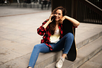 Fototapeta premium Portrait of young beautiful girl with skateboard. Happy smiling woman talking to the phone.