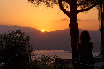 Orangener Sonnenuntergang hinter Bergen mit Baum und Frau im Vordergrund, Capoliveri, Elba, Italien