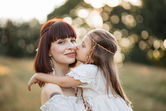 Close up of lovely little 4-years old girl, embracing her pretty caring mom, and kissing her in cheek. Indian family outdoors in the field. Boho woman with child walking at summer sunset