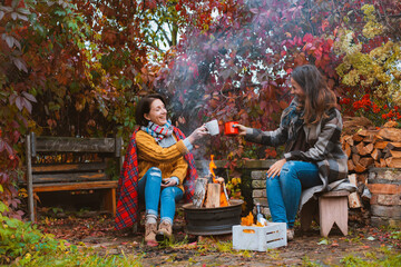 two happy joyful best friends sitting outside in autumn garden by campfire with bottle of red wine